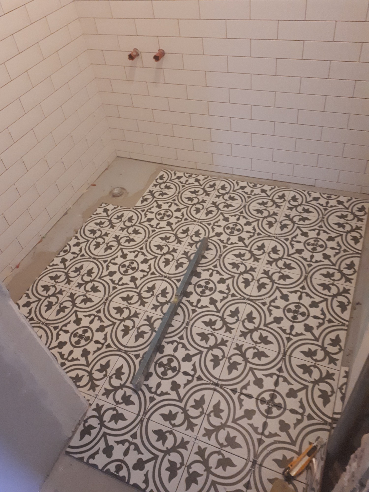 Układanie dekoracyjnych, czarno-białych płytek podłogowych w łazience z białymi, prostokątnymi płytkami ściennymi. Widoczna poziomica i nóż do cięcia płytek.