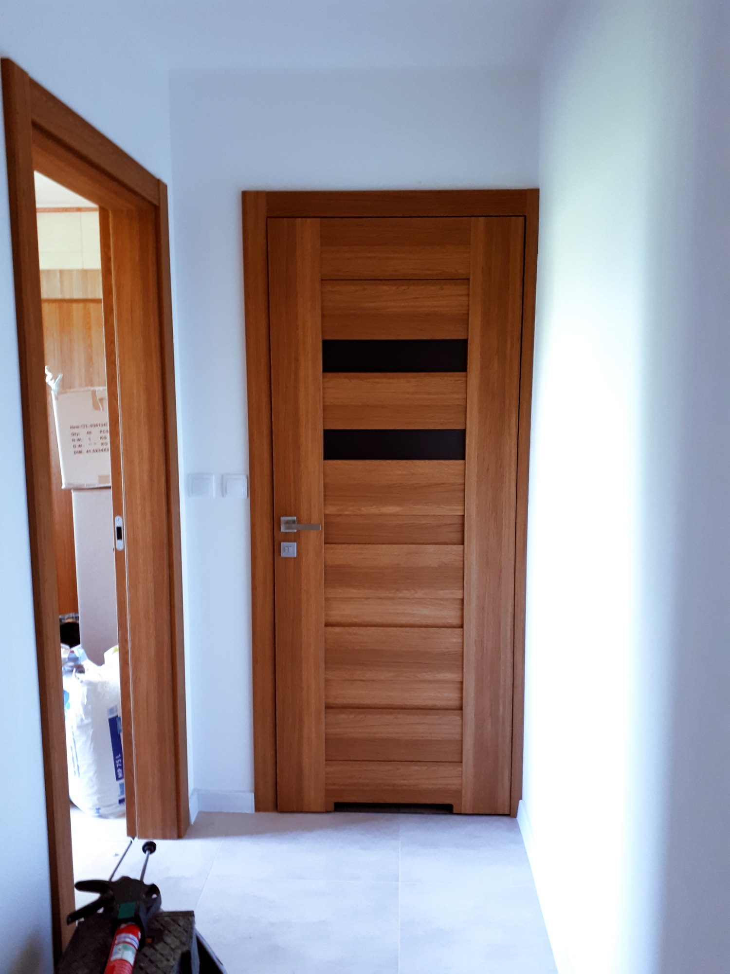 Nowoczesne, drewniane drzwi wewnętrzne z czarnymi akcentami, zamontowane w białym korytarzu. Widoczne narzędzia i materiały budowlane w tle.