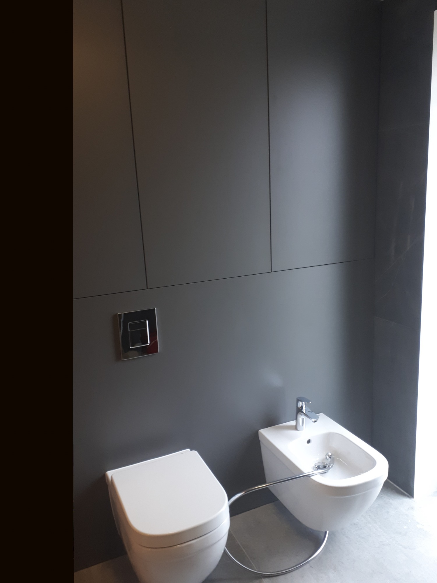 Nowoczesna łazienka z szarą zabudową, wiszącą toaletą i bidetem z chromowaną armaturą. Minimalistyczny design, gładkie powierzchnie, subtelne oświetlenie.