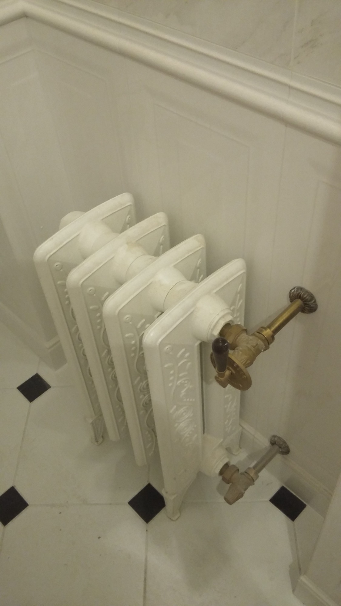 Biały, dekoracyjny grzejnik żeberkowy z mosiężnymi zaworami w łazience z białą boazerią i podłogą w biało-czarną szachownicę. Ujęcie z góry, blisko ściany.