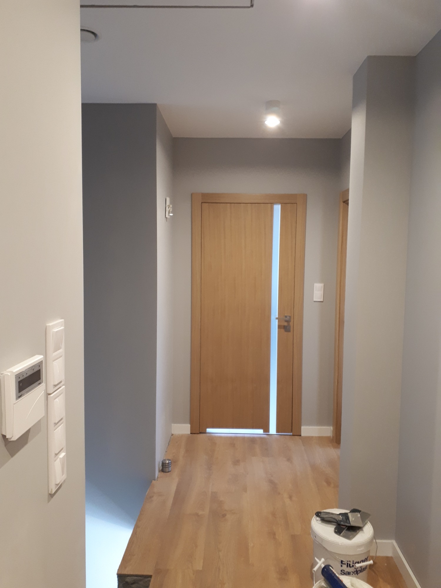 Jasne wnętrze po remoncie: szare ściany, drewniana podłoga i drzwi z przeszkleniem. Wiadro z narzędziami malarskimi w rogu sugeruje świeże wykończenie.