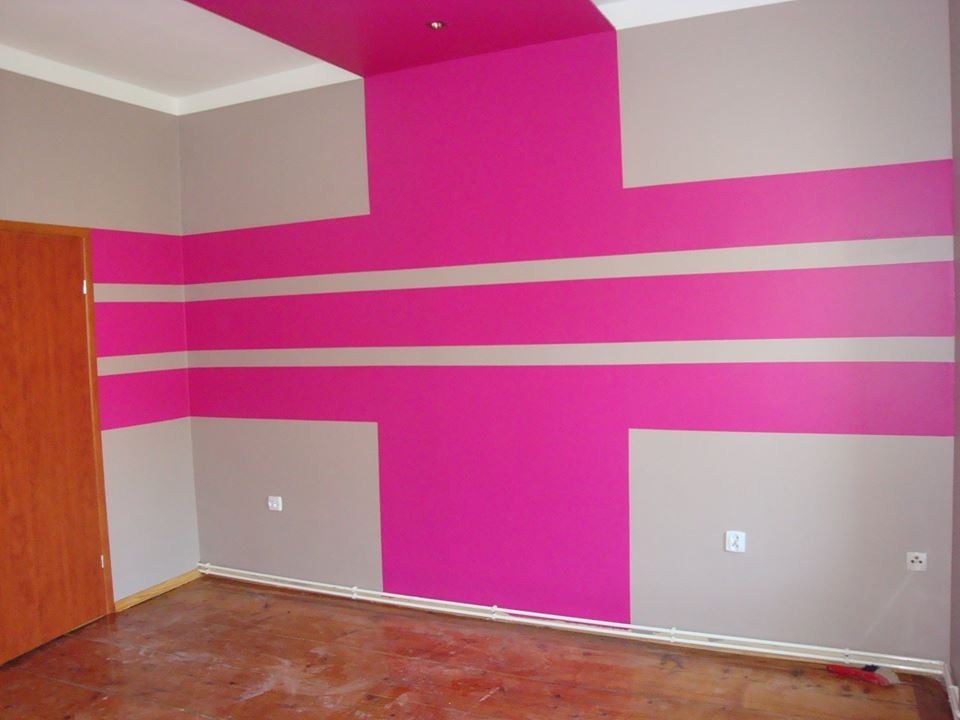 Pokój po malowaniu w geometryczne wzory: różowe pasy i bloki na szarym tle. Widoczna drewniana podłoga i fragment drzwi. Nietypowe połączenie kolorów.