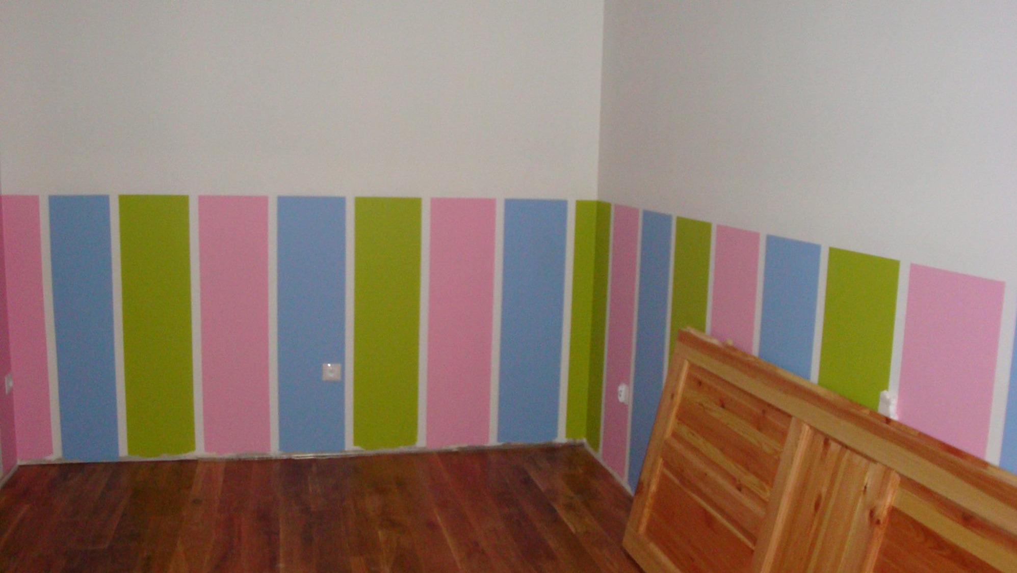 Pokój z pomalowanymi ścianami w pionowe pasy: różowe, zielone i niebieskie. Podłoga drewniana, w rogu fragment drewnianych drzwi. Widoczne gniazdka elektryczne.