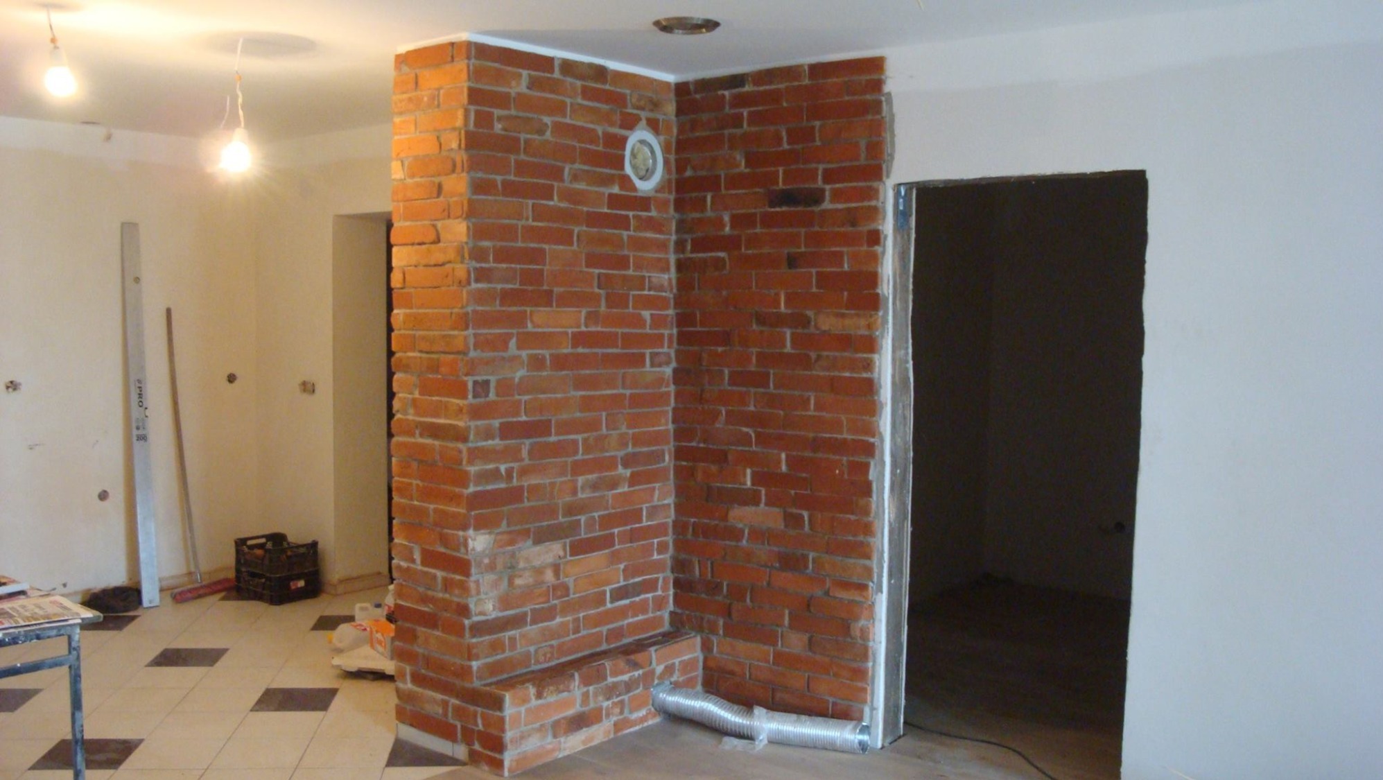 Wykończenie wnętrza: narożny fragment ściany z czerwonej cegły z widoczną wentylacją, surowy otwór drzwiowy i fragment podłogi w trakcie remontu.