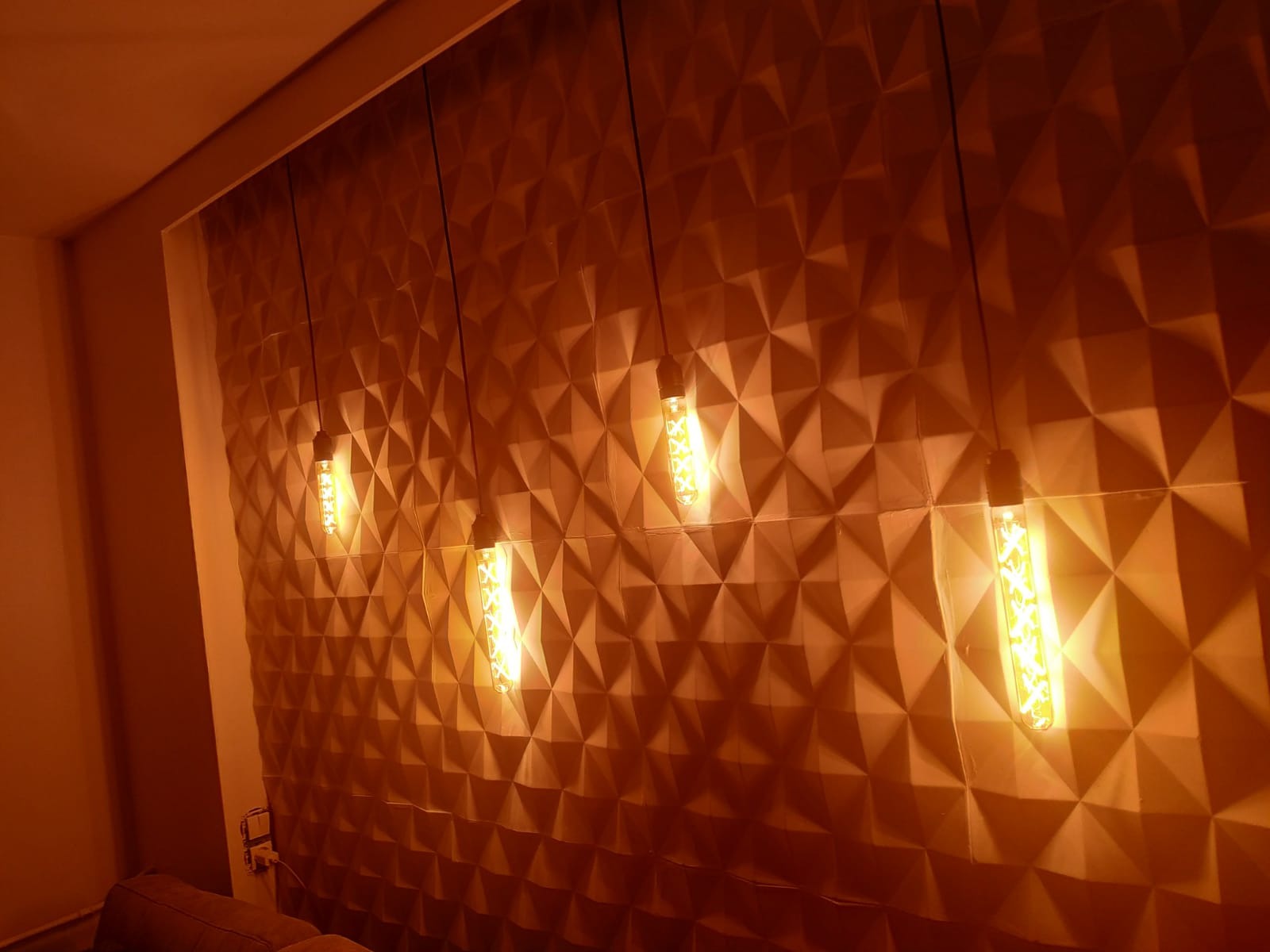 Ściana z trójwymiarowych paneli w ciepłym odcieniu, oświetlona czterema wiszącymi żarówkami w stylu retro, tworzącymi geometryczny wzór światła i cienia.