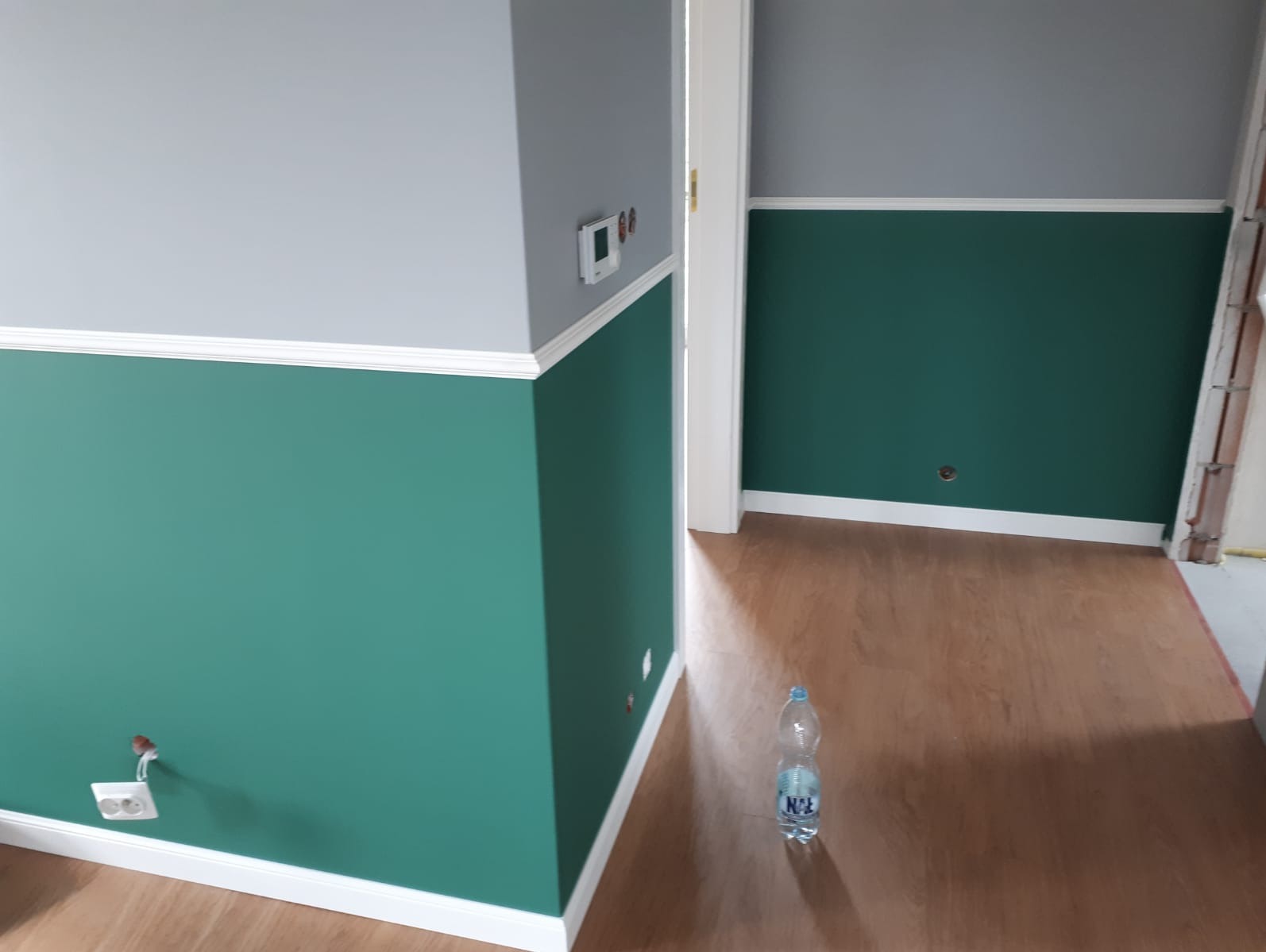 Pomieszczenie po malowaniu ścian na szaro i zielono, z białymi listwami przypodłogowymi i ozdobnymi. Na podłodze butelka wody.