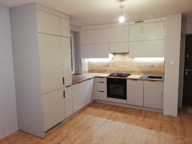 Jasna kuchnia z drewnianym blatem i panelami. Minimalistyczne szafki bez uchwytów, oświetlenie LED pod szafkami wiszącymi. Nowoczesny design.