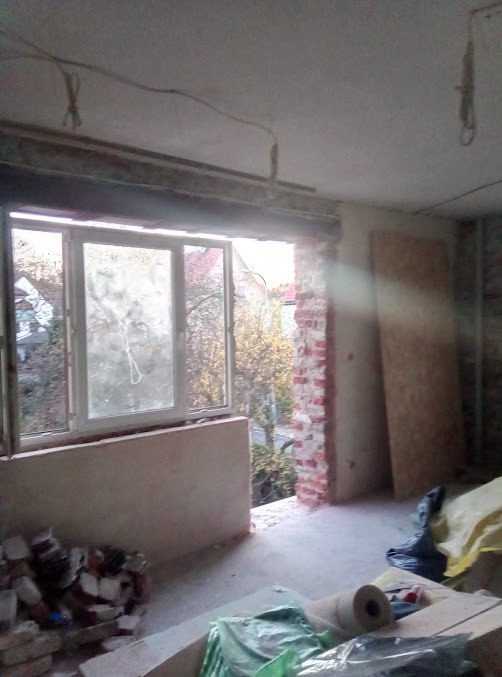 Wnętrze w trakcie remontu: okno balkonowe z widokiem na zewnątrz, odsłonięta cegła, materiały budowlane na podłodze. Widoczne kable wystające z sufitu.