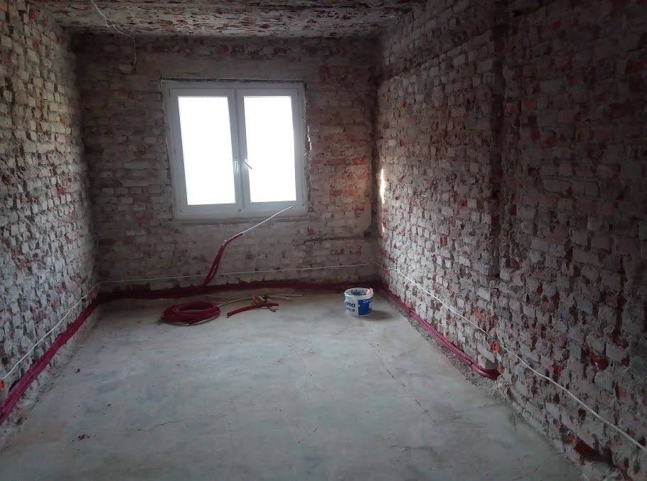Pokój w trakcie remontu: surowe ceglane ściany, okno, instalacje w listwach przypodłogowych, wiadro farby i narzędzia na szarym podłożu. Widok na wnętrze przed wykończeniem.