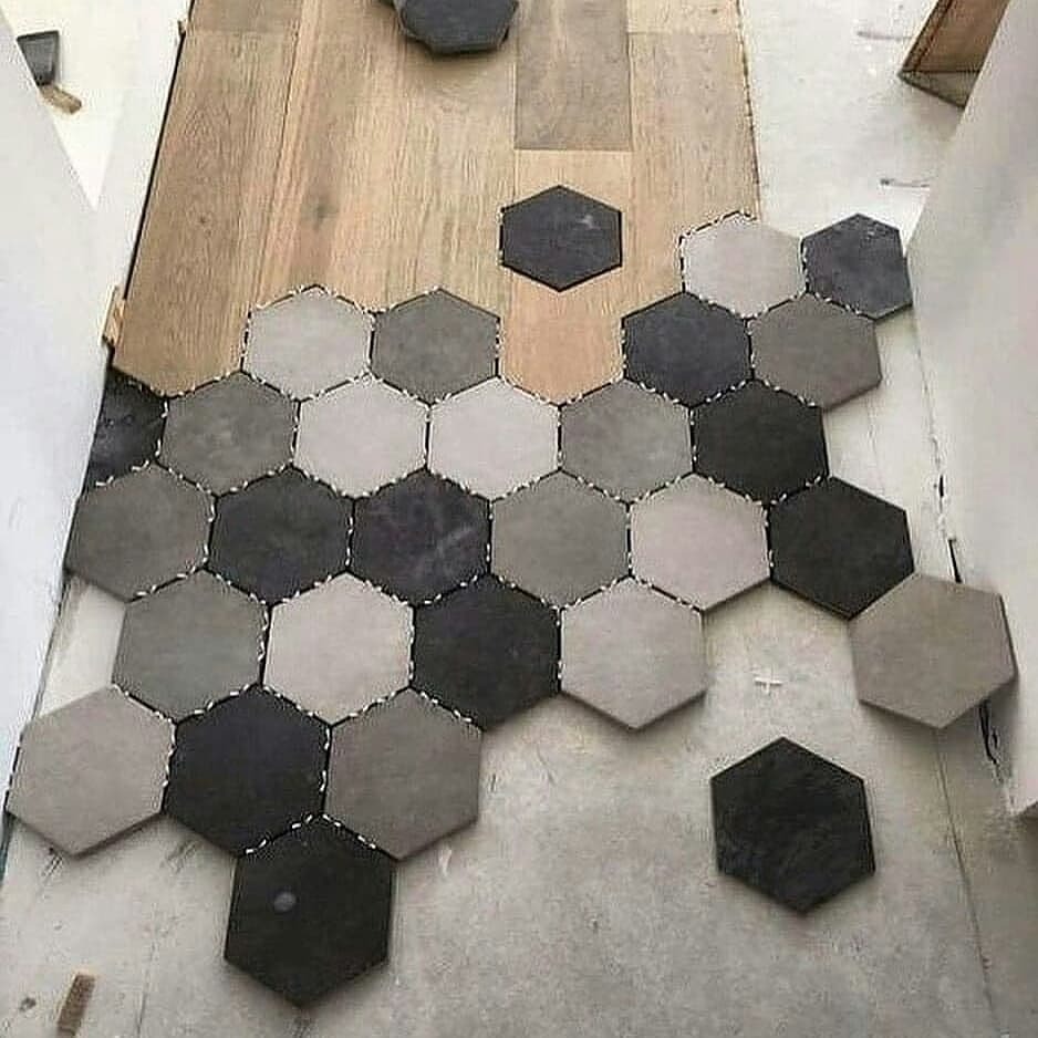 Układanie heksagonalnych płytek w odcieniach szarości na podłodze, widoczne fugi i fragmenty drewnianej podłogi. Nietypowy wzór i kolorystyka.