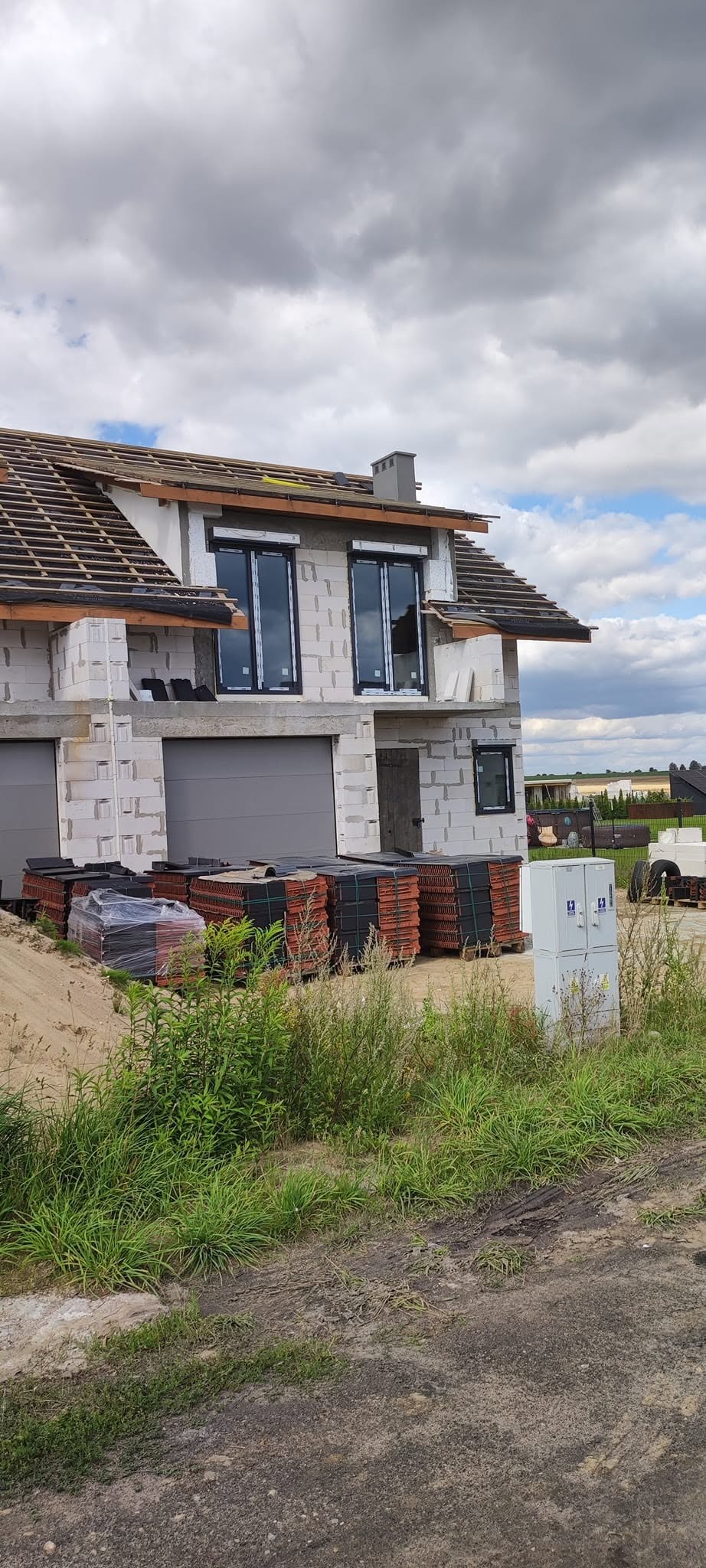 Surowy dom z pustaków, z oknami i dachem w budowie. Na ziemi palety z dachówkami. Widoczne elementy konstrukcyjne i materiały budowlane. Etap prac dekarskich.