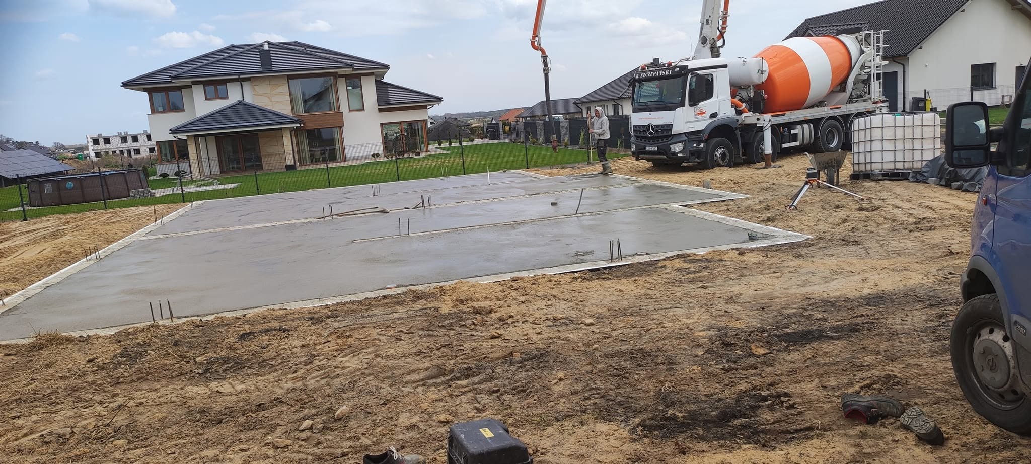 Wylany fundament pod dom jednorodzinny z widocznym zbrojeniem. Na drugim planie betoniarka i budynek w trakcie budowy. Prace ziemne w toku.