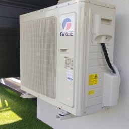 Sevro klimatyzacja, pompy ciepła - Zewnętrzna jednostka klimatyzacji Gree zamontowana na ścianie budynku z widocznymi przewodami i elementami mocującymi, na tle sztucznej trawy.