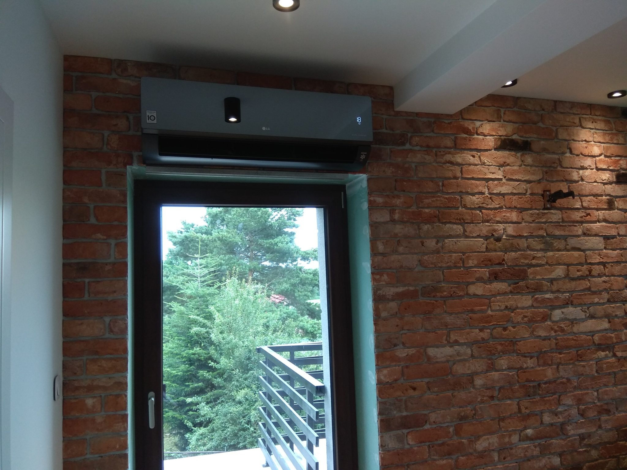 Szary klimatyzator LG zamontowany na ceglanej ścianie nad drzwiami balkonowymi z widokiem na zieleń. Widoczne oświetlenie sufitowe.