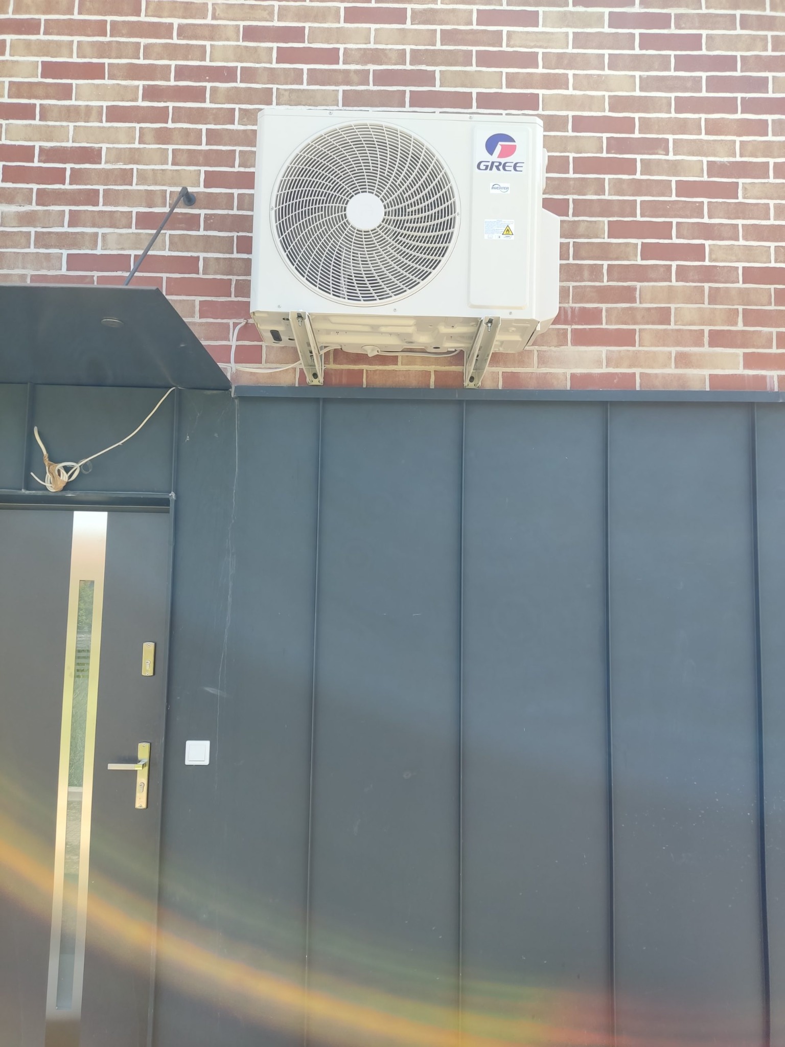 Biały klimatyzator marki Gree zamontowany na ceglanej ścianie nad szarą, metalową bramą. Widoczne przewody i elementy montażowe.