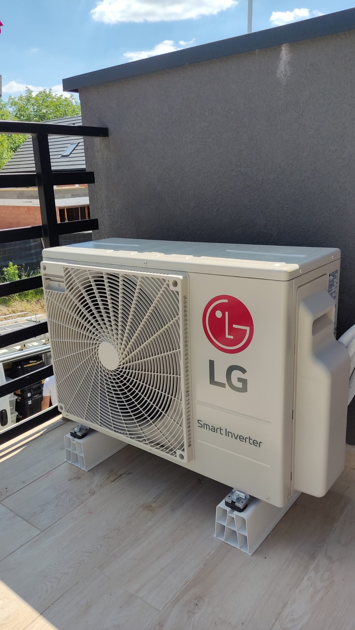Zewnętrzna jednostka klimatyzacji LG Smart Inverter zamontowana na balkonie, widoczna z boku, z elementami mocującymi. Tło stanowi szara ściana budynku i fragment balustrady.