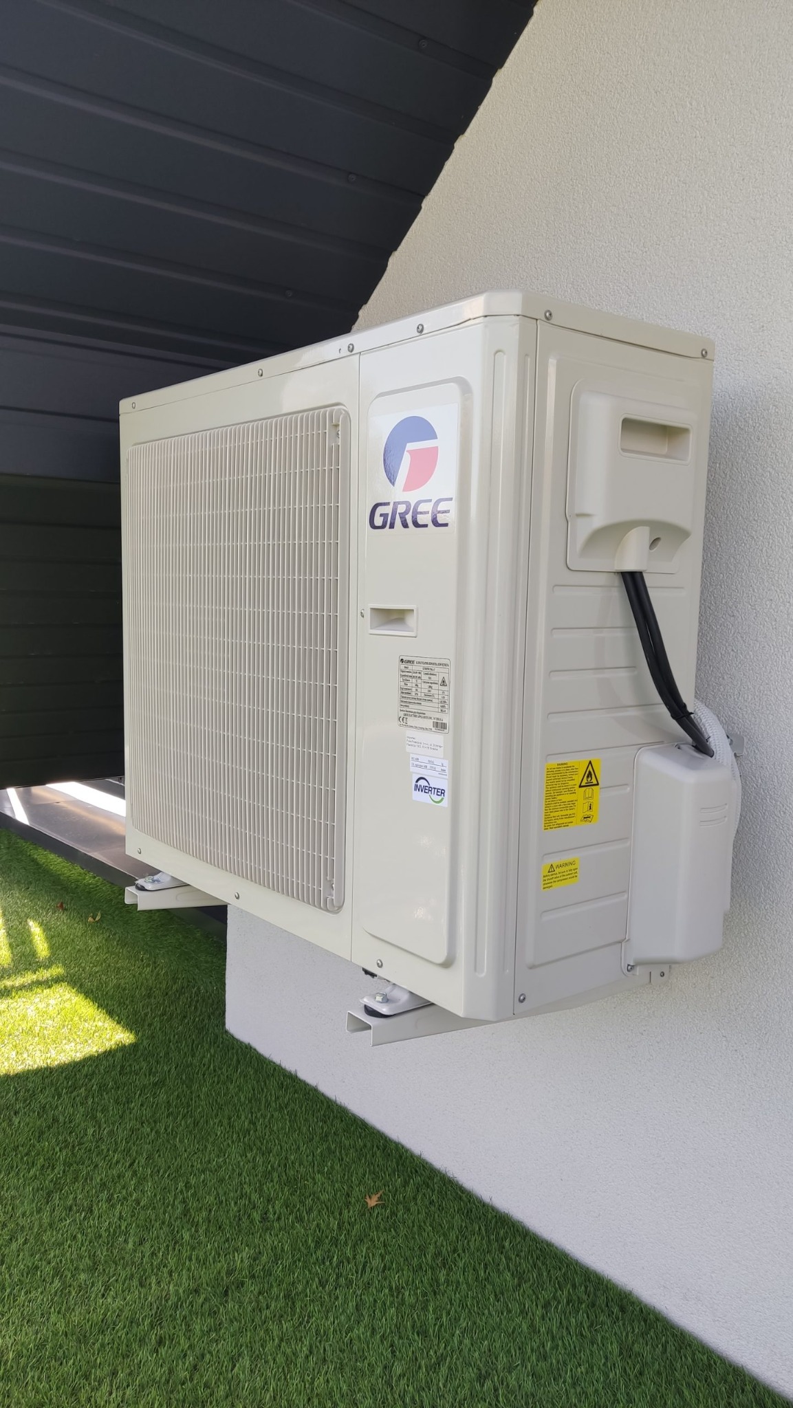 Zewnętrzna jednostka klimatyzacji Gree zamontowana na ścianie budynku z widocznymi przewodami i elementami mocującymi, na tle sztucznej trawy.