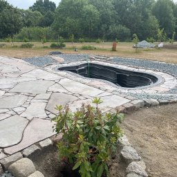 MATI Mateusz Dering - Oczko wodne z czarną formą, otoczone szarym żwirem i kamienną obudową. W tle widoczny ogród z drzewami i rabatami. Kamienny chodnik z nieregularnych płyt.