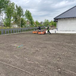 MATI Mateusz Dering - Przygotowanie gruntu pod trawnik: dwóch mężczyzn używa walców spalinowych na dużej działce przy nowoczesnym domu. Widoczne ślady walcowania i fragment niebieskiego węża.