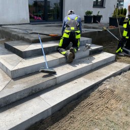 MATI Mateusz Dering - Montaż schodów zewnętrznych z szarych płyt kamiennych. Dwóch pracowników w odzieży roboczej układa stopnie. Widoczne narzędzia: młotek, poziomica, szczotka.