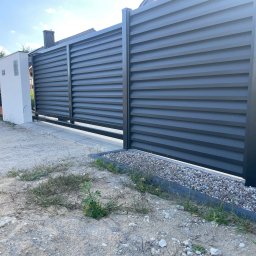 MATI Mateusz Dering - Nowoczesne, poziome, szare ogrodzenie panelowe z przesuwną bramą. Widoczny fragment posesji i osoba przy słupku. Uwagę zwraca minimalistyczny design i surowy charakter otoczenia.