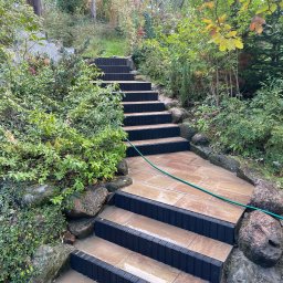 MATI Mateusz Dering - Kamienne schody ogrodowe z czarnymi obrzeżami, prowadzące w górę przez bujną roślinność. Wąż ogrodowy leży na schodach, sugerując niedawne czyszczenie.