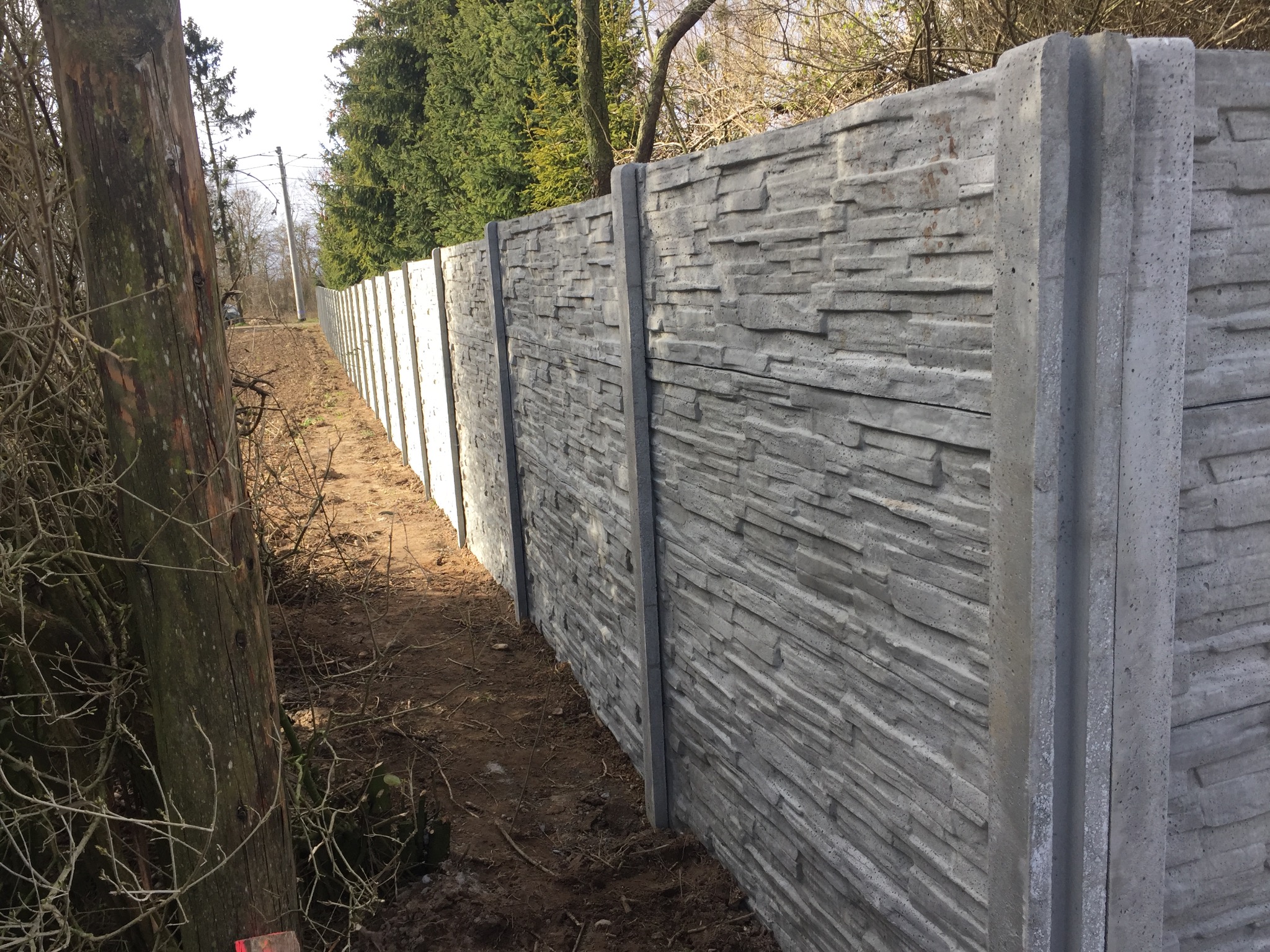 Długi, betonowy płot o fakturze kamienia, oddzielający posesję. Widoczne słupki i panele. Tło stanowią drzewa i krzewy. Ujęcie z niskiej perspektywy.