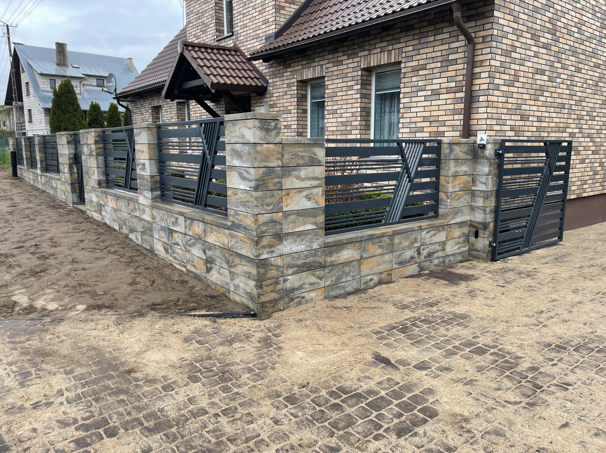 Nowoczesne ogrodzenie z szarego kamienia i metalu, przylegające do domu z cegły. Brama wjazdowa z metalowymi przęsłami. Kostka brukowa na podjeździe.