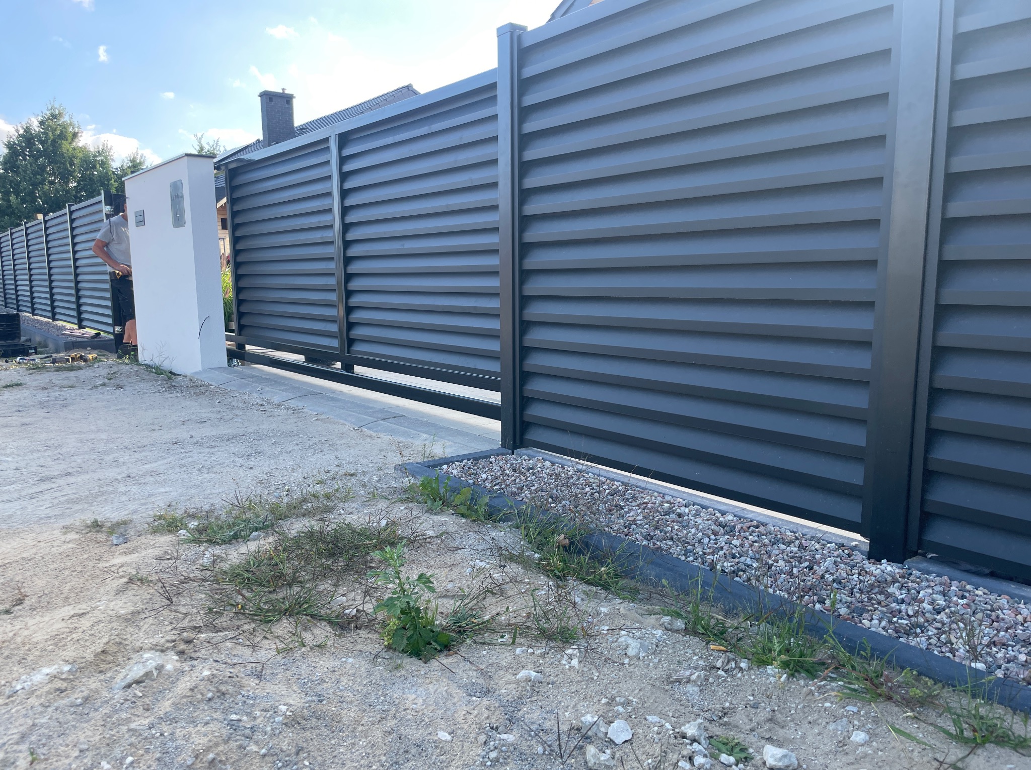 Nowoczesne, poziome, szare ogrodzenie panelowe z przesuwną bramą. Widoczny fragment posesji i osoba przy słupku. Uwagę zwraca minimalistyczny design i surowy charakter otoczenia.