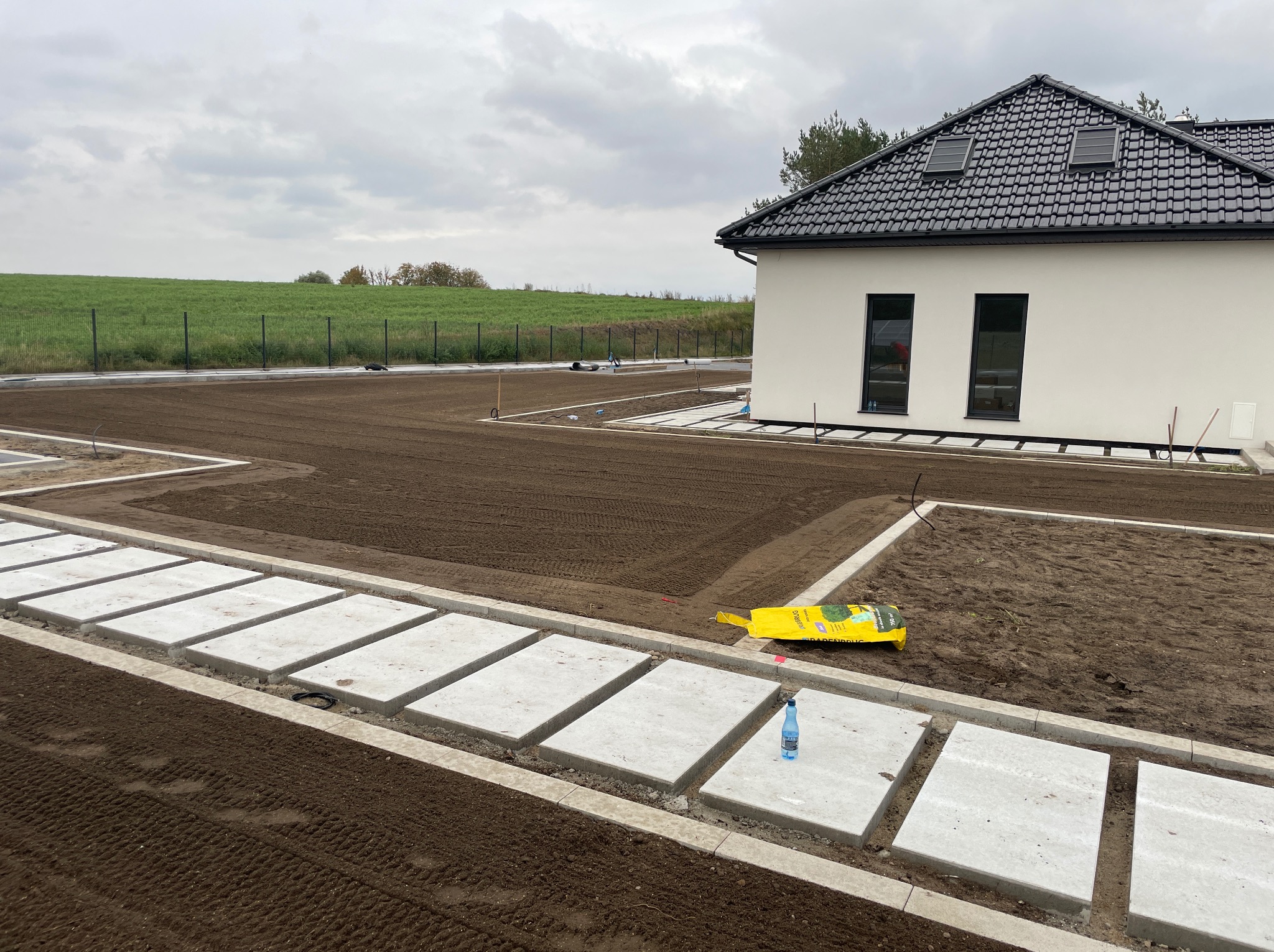 Przygotowany teren pod trawnik z ułożonymi płytami chodnikowymi i obrzeżami, widok na nowoczesny dom z czarną dachówką i zielone pole w tle.