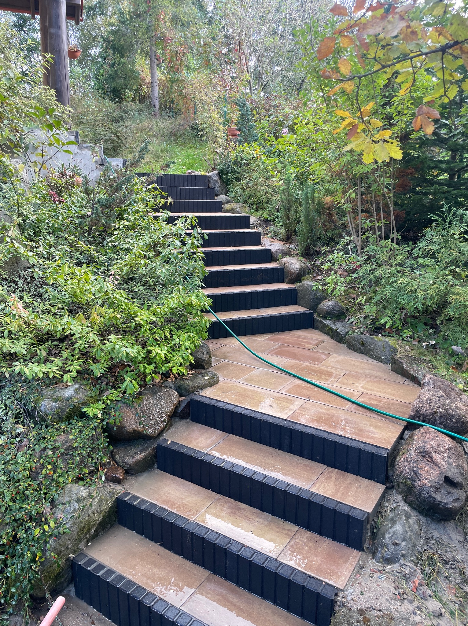 Kamienne schody ogrodowe z czarnymi obrzeżami, prowadzące w górę przez bujną roślinność. Wąż ogrodowy leży na schodach, sugerując niedawne czyszczenie.