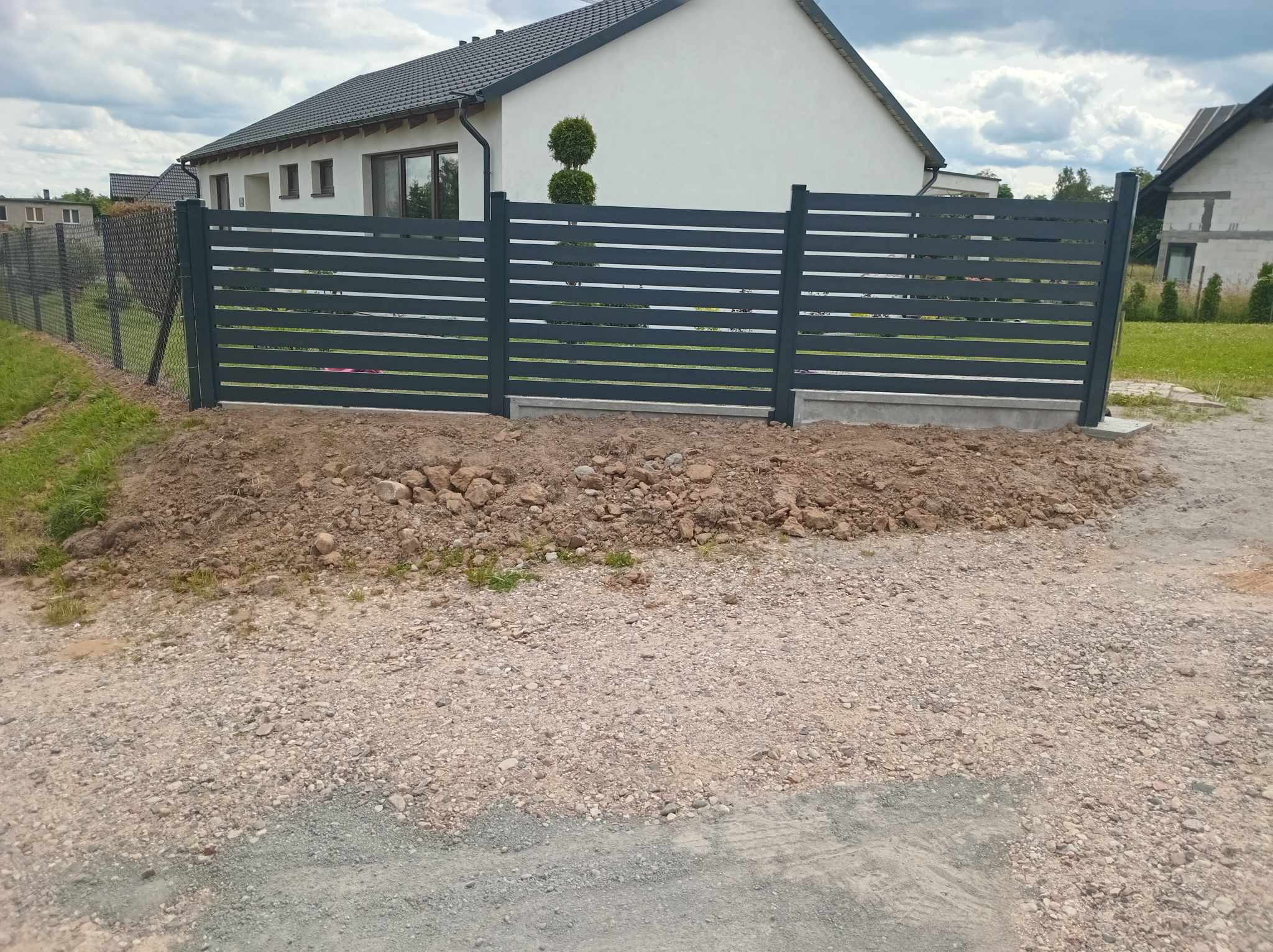 Nowoczesne, poziome, szare ogrodzenie panelowe z betonową podmurówką, na tle domu z szarą dachówką i nieba. Widoczny teren przygotowany pod dalsze prace.