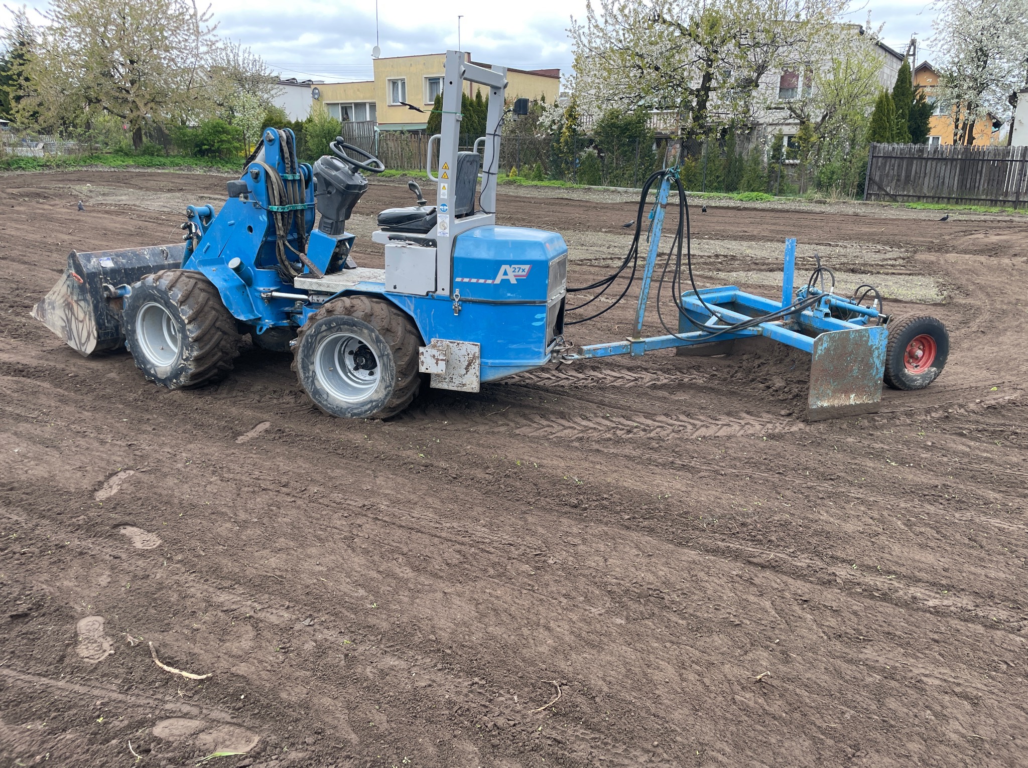 Niebieski traktor z równiarką na przygotowanym polu pod zasiew, w tle zabudowa jednorodzinna i kwitnące drzewa. Widoczne ślady opon na ziemi.