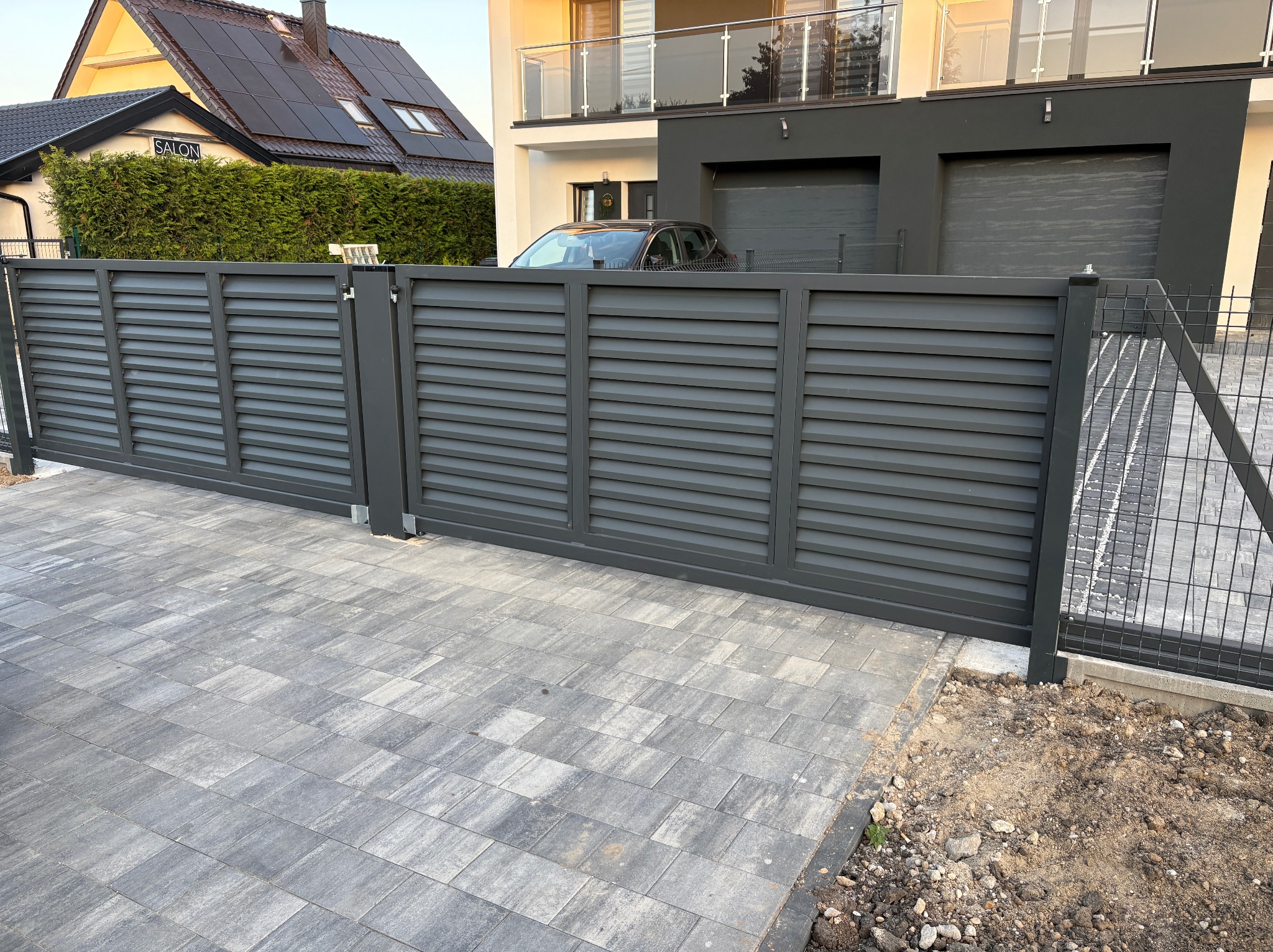 Nowoczesna, przesuwna brama lamelowa w kolorze grafitowym, zamontowana na posesji z kostki brukowej. W tle dom z panelami słonecznymi i salon fryzjerski.