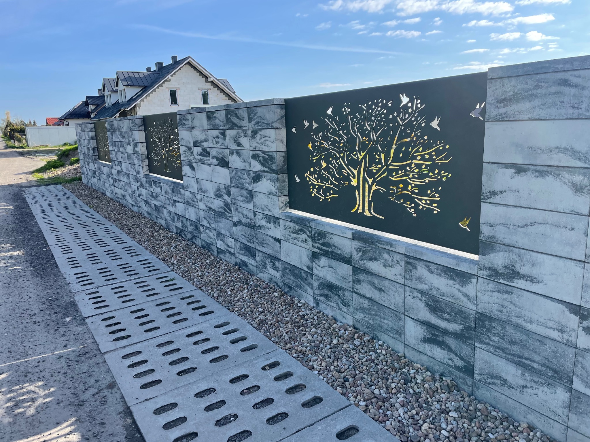Nowoczesny, modułowy płot z szarego betonu architektonicznego, z dekoracyjnymi panelami z motywem drzewa i ptaków. U podstawy płotu wysypane drobne kamienie.