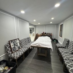 Fajna Budowa Dmytro Kobiuk - Szare panele podłogowe w pokoju z listwami ściennymi. Stół przykryty płachtą, krzesła z szarą tapicerką w biały wzór. W tle schody i wyposażenie.