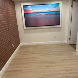 Fajna Budowa Dmytro Kobiuk - Wykończone wnętrze z panelami podłogowymi, ceglaną ścianą i telewizorem w białej ramie, prezentującym widok plaży o zachodzie słońca. Nowoczesne oświetlenie sufitowe.