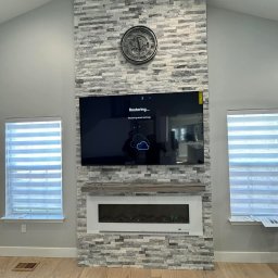 Fajna Budowa Dmytro Kobiuk - Ściana z dekoracyjnej szarej cegły z kominkiem elektrycznym i telewizorem. Na górze zegar. Parkiet na podłodze. Okna z żaluzjami po bokach.