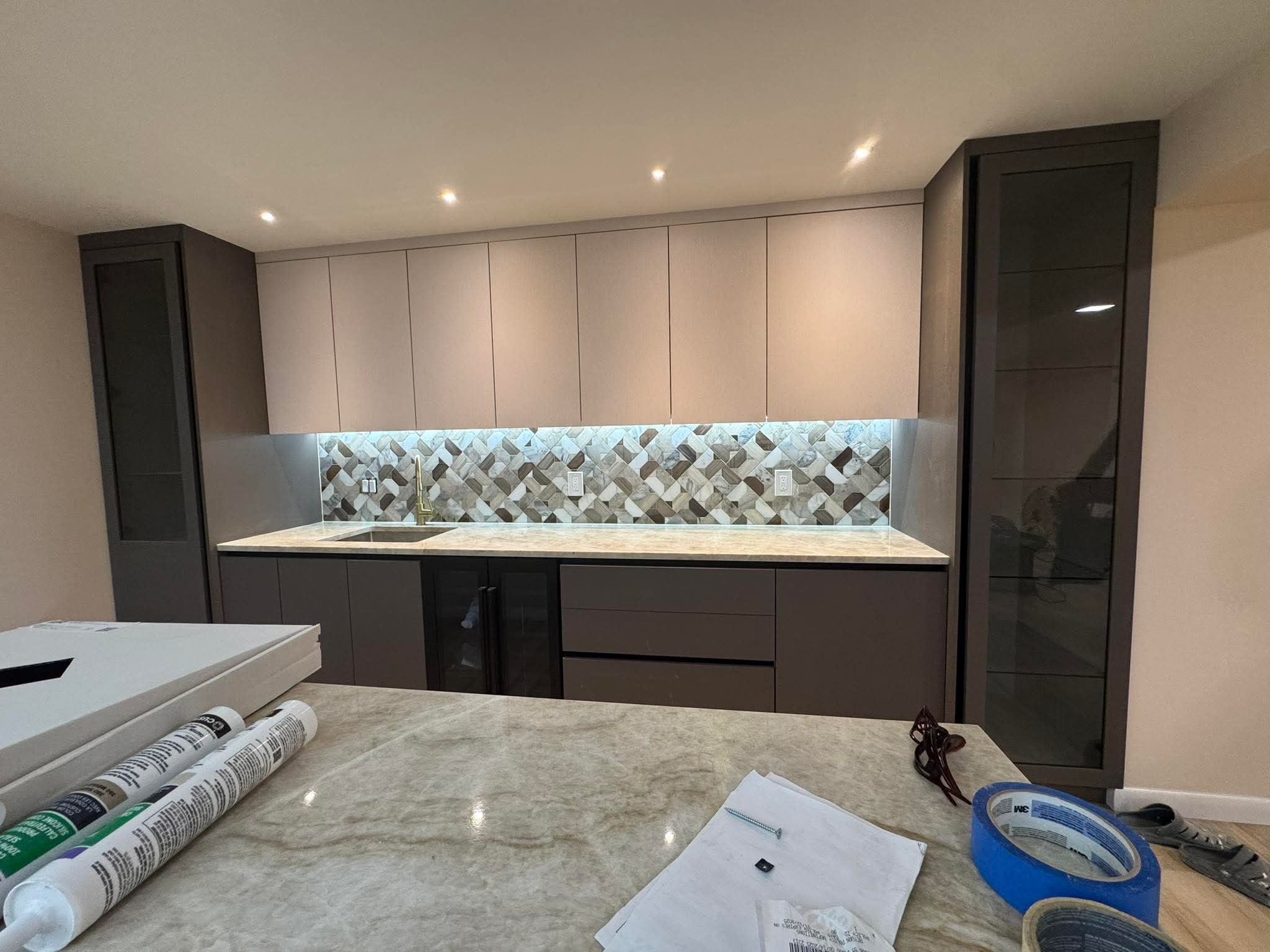 Nowoczesna kuchnia z mozaiką nad blatem, oświetleniem LED i szarymi szafkami. Na pierwszym planie materiały montażowe na blacie.