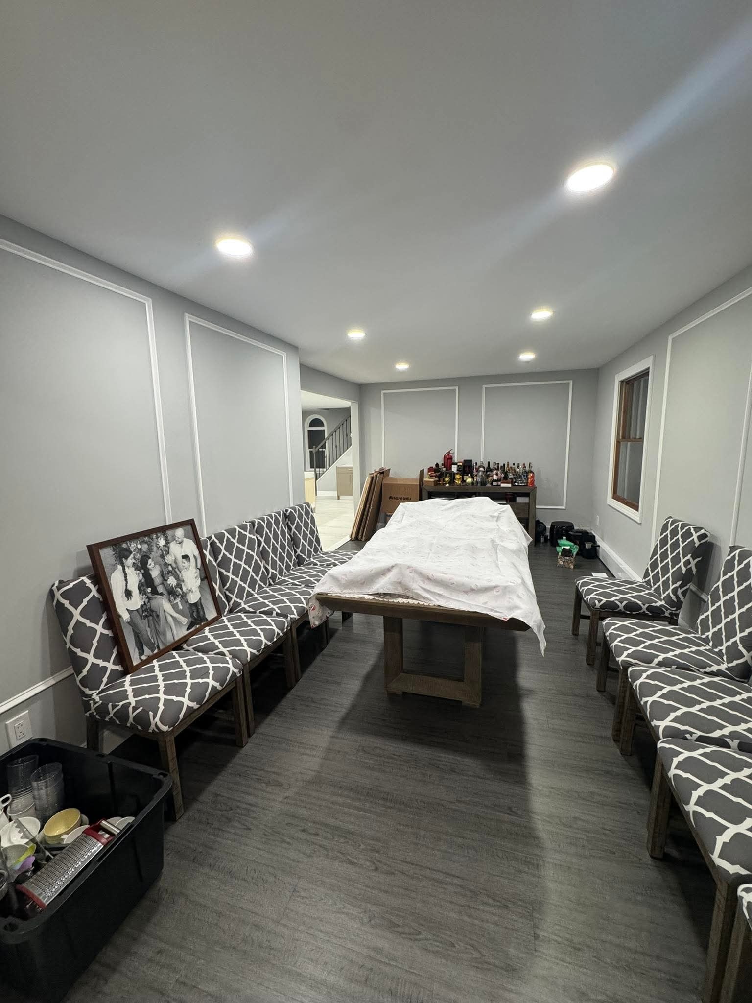 Szare panele podłogowe w pokoju z listwami ściennymi. Stół przykryty płachtą, krzesła z szarą tapicerką w biały wzór. W tle schody i wyposażenie.