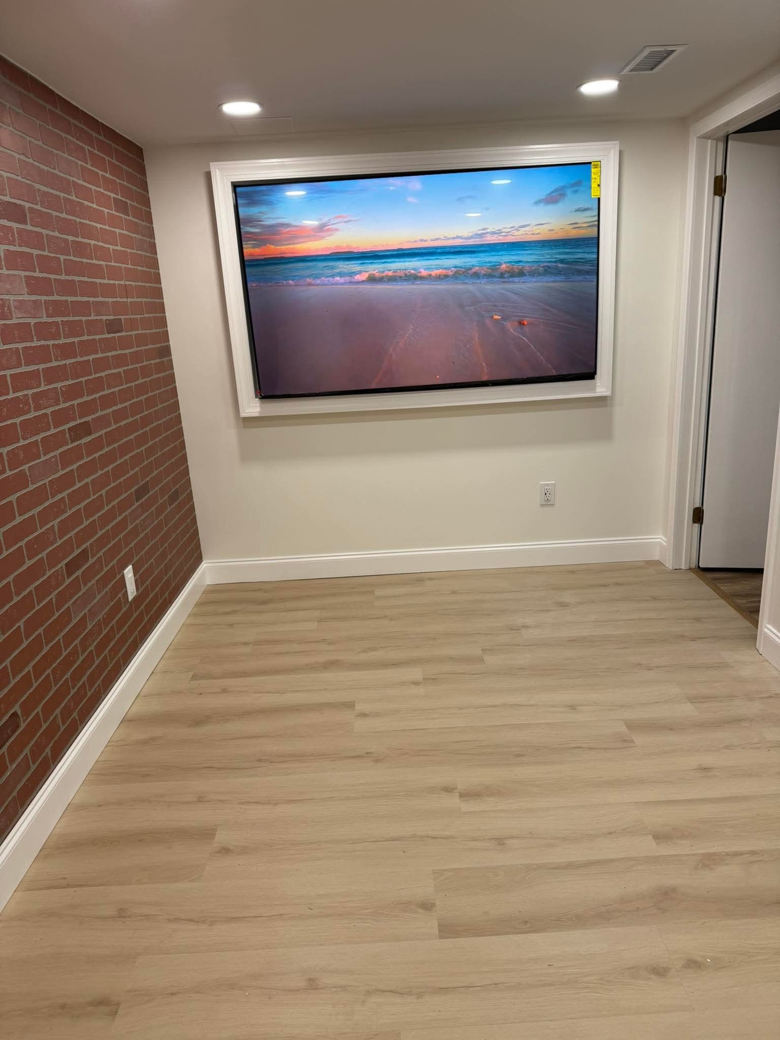 Wykończone wnętrze z panelami podłogowymi, ceglaną ścianą i telewizorem w białej ramie, prezentującym widok plaży o zachodzie słońca. Nowoczesne oświetlenie sufitowe.