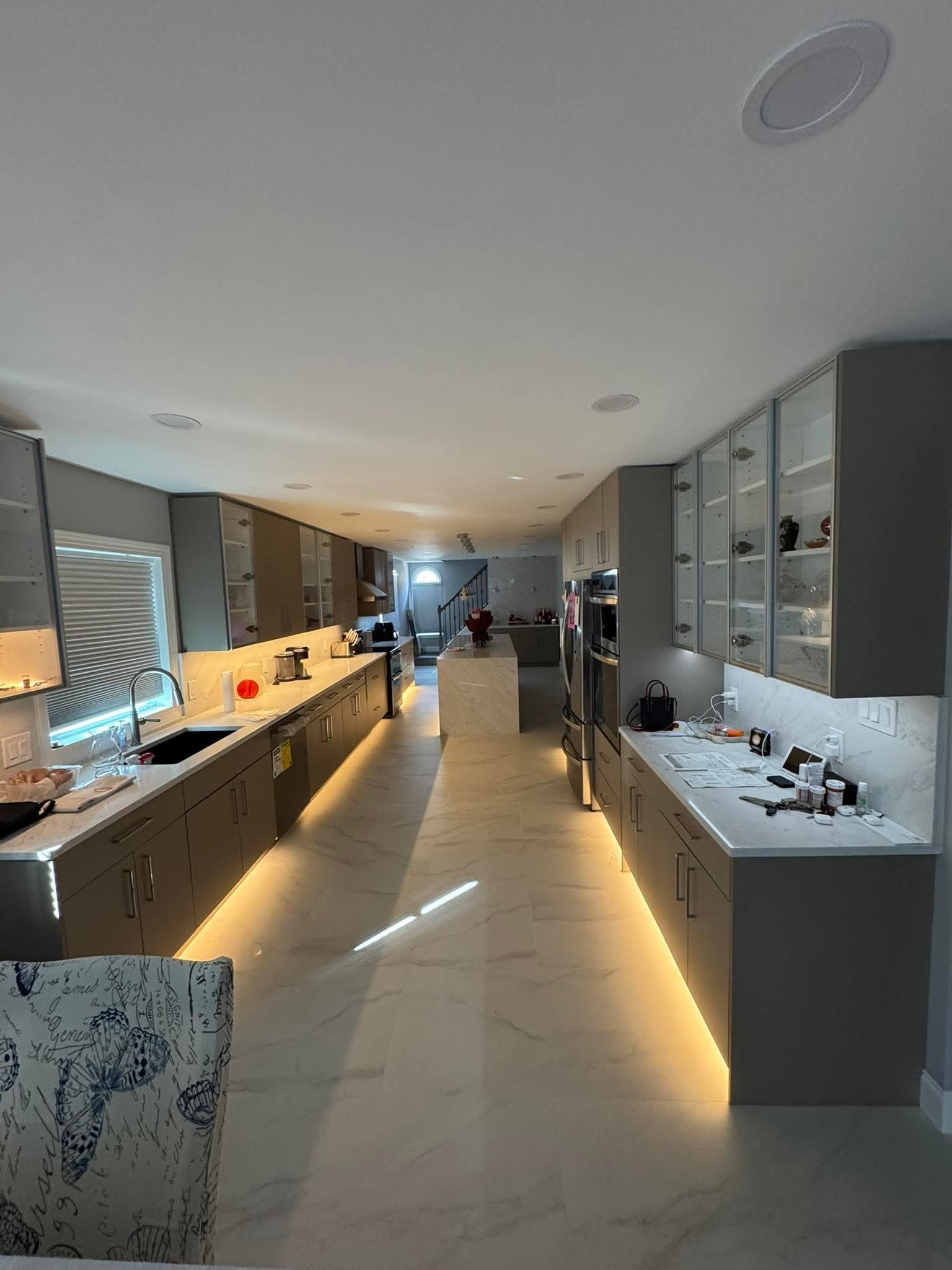 Długa, jasna kuchnia z szarymi szafkami, marmurowymi blatami i podświetleniem LED pod szafkami. Widok w głąb pomieszczenia, w tle wyspa kuchenna i schody.