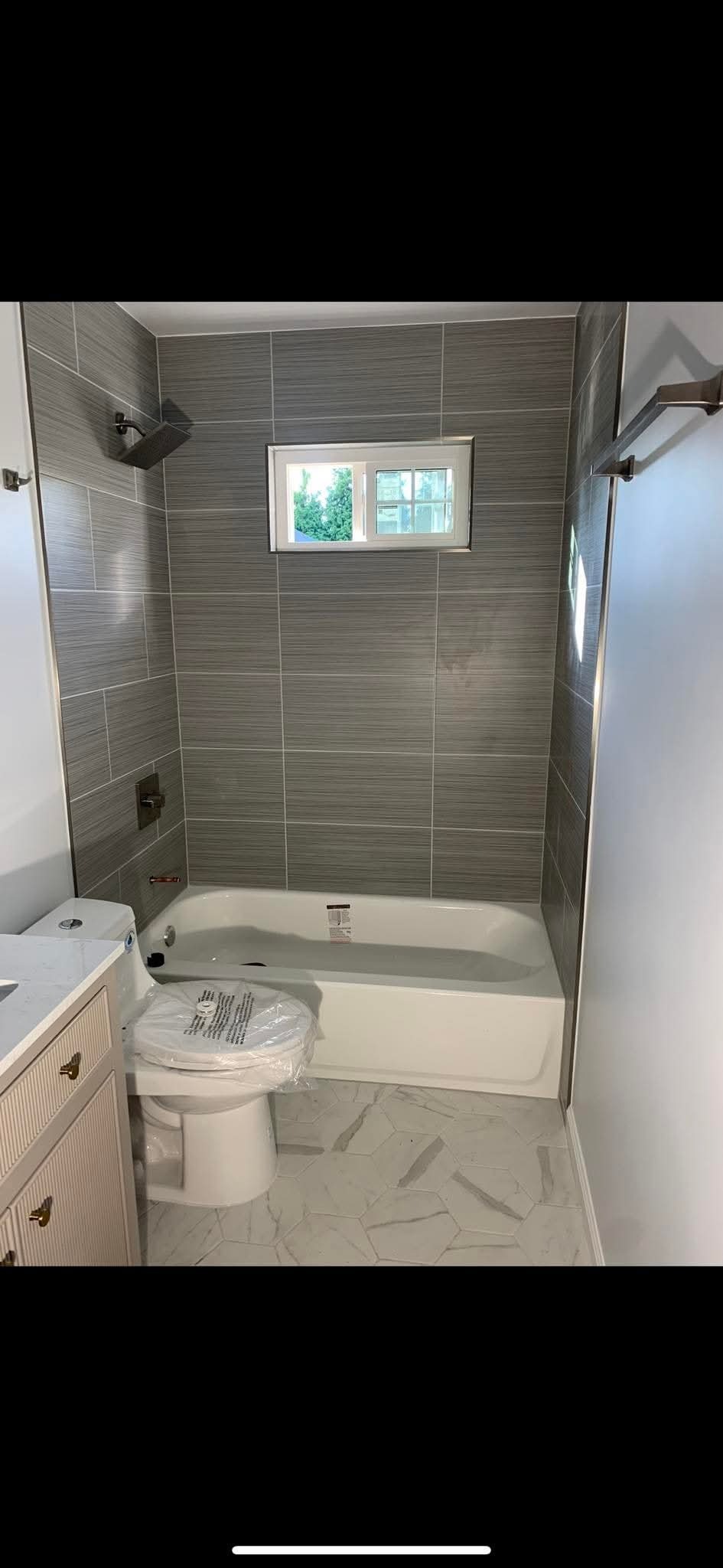 Wykończona łazienka z szarymi płytkami imitującymi drewno, białą wanną i toaletą z folią ochronną. Małe okno z widokiem na zieleń. Marmurowe heksagonalne płytki podłogowe.