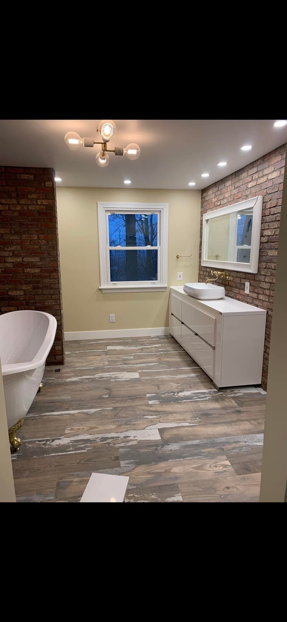 Ukończona łazienka z ceglaną ścianą, drewnopodobną podłogą i białą szafką z umywalką. Widoczne okno i nowoczesne oświetlenie sufitowe.