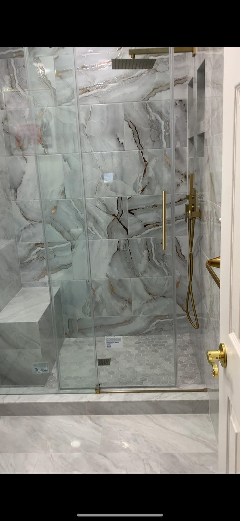 Wykończona kabina prysznicowa z marmurowymi płytkami w odcieniach szarości i złota armatura. Widoczna szklana ściana, siedzisko i mozaika na podłodze.