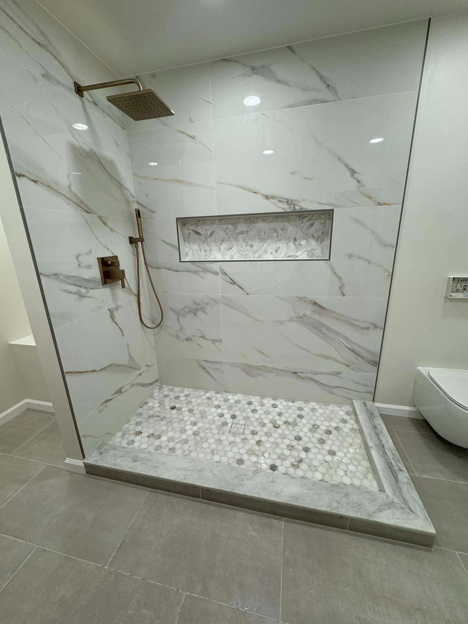 Wykończona kabina prysznicowa z marmurowymi płytkami imitującymi naturalny kamień, mozaiką na podłodze i wnęką dekoracyjną. Armatura w kolorze starego złota.