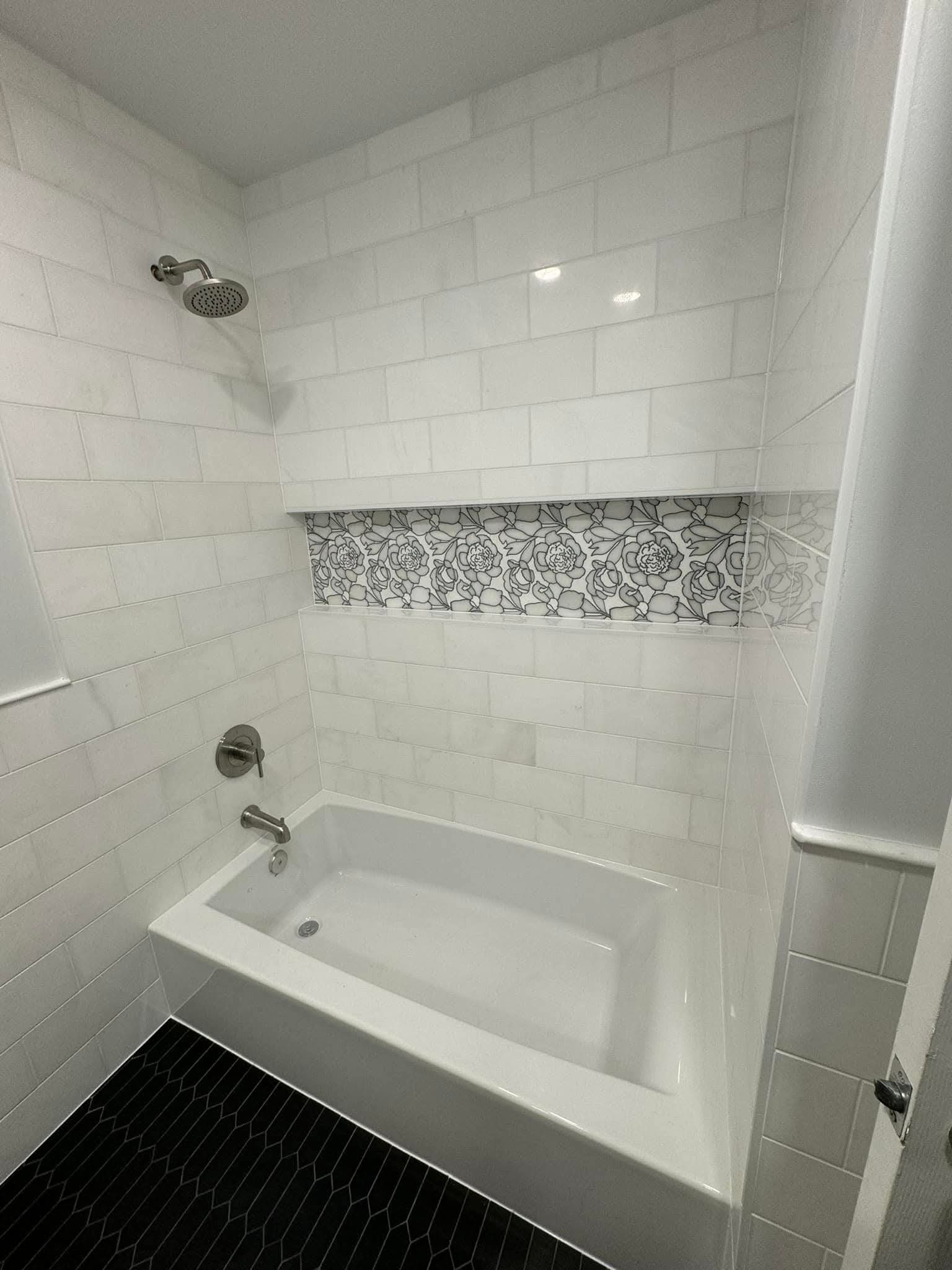 Biała wanna obudowana płytkami z dekoracyjnym pasem kwiatowym. Czarne, wąskie płytki podłogowe ułożone w jodełkę. Nowoczesna armatura łazienkowa.