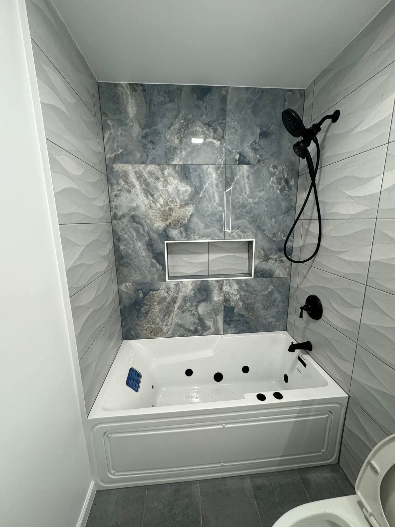 Wykończona łazienka z wanną jacuzzi, czarną armaturą i szarymi płytkami imitującymi fale oraz marmur. W ścianie wnęka wykończona płytkami. Widok na fragment toalety.