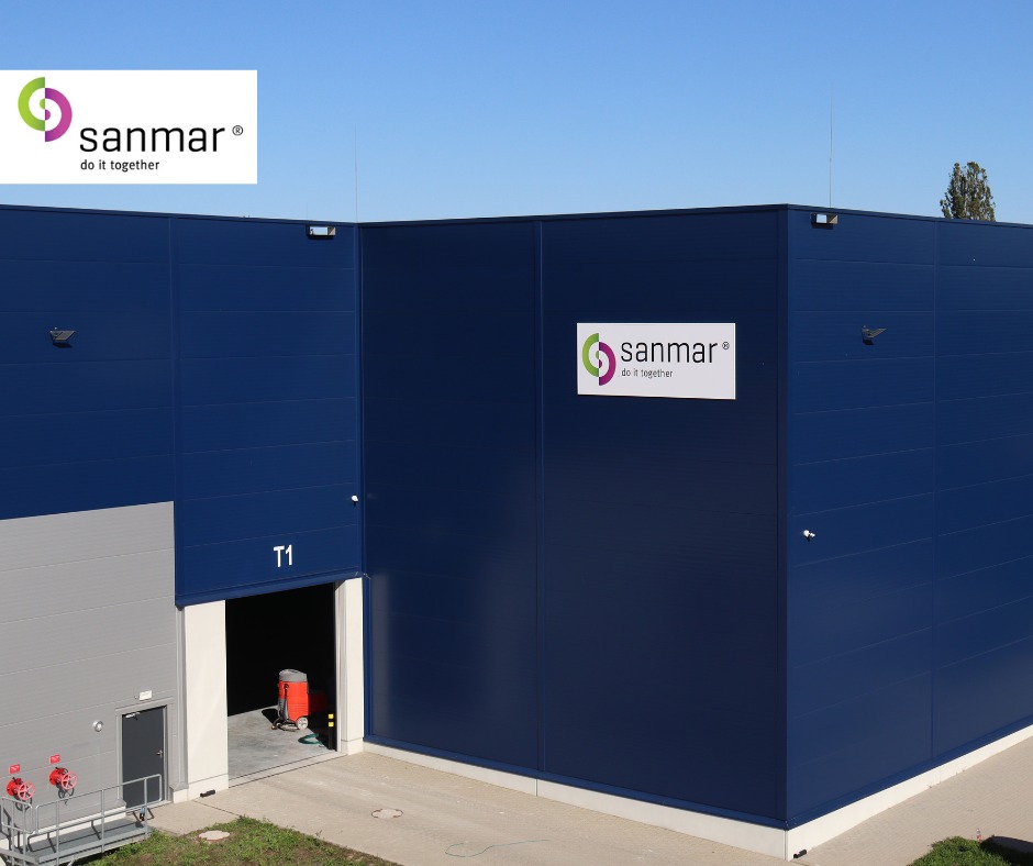 Nowoczesny magazyn w kolorze granatowym z logo Sanmar, widoczny otwarty wjazd T1 i fragment zieleni. Budynek o prostej architekturze, oświetlony naturalnym światłem.