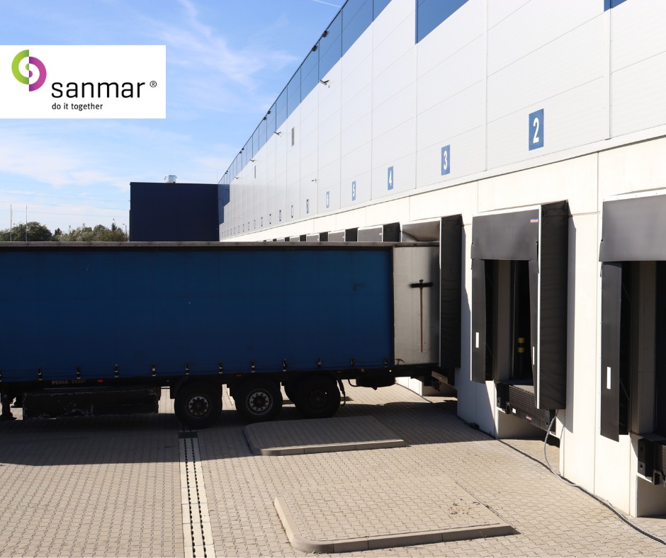 Ciężarówka z naczepą przy rampie magazynowej z numerami. Błękitne niebo w tle. Widoczny fragment logo firmy Sanmar.